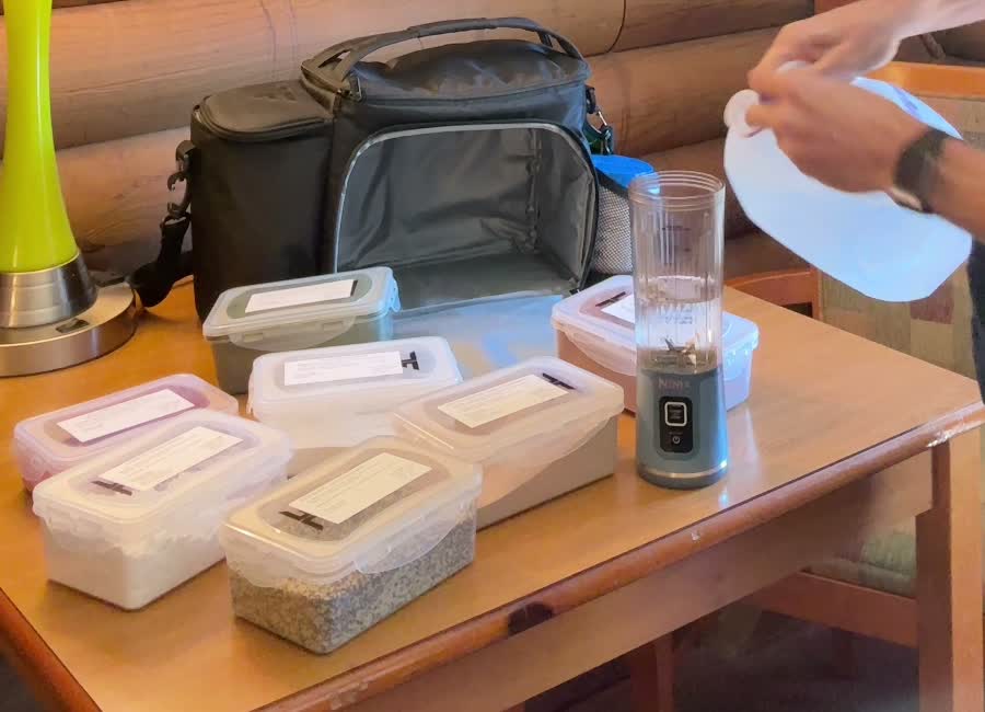 Labeled ingredient containers, a portable blender, and an open meal-prep backpack on the hotel table